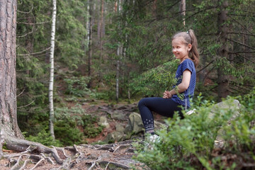 Naklejka na ściany i meble The girl in a purple T-shirt in the woods with a sprig of blueberries sat on a stone and smiles