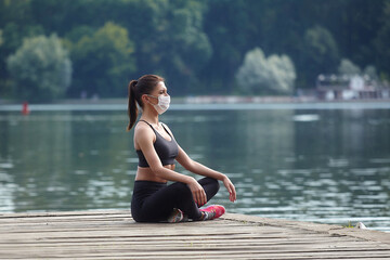 Attractive girl in a mask and sportswear is doing gymnastic exercises on a wooden pier in a city park. safe outdoor workout concept