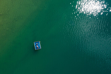 Floating platform with a sun tanning couple aerial top view. Bright sn reflecting in green lake waves. Peruca Lake, Dalmatia, Croatia - obrazy, fototapety, plakaty