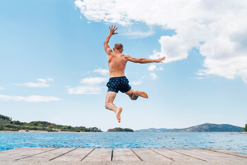 Middle-aged man dressed swimming trunks funny jumping to the waves from the boat pier as he having...