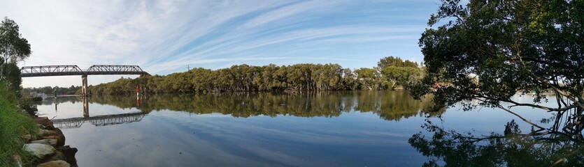 Beautiful panoramic view of a river with reflections of tall pedestrian bridge, trees and blue sky, Parramatta river, Rydalmere, New South Wales, Australia