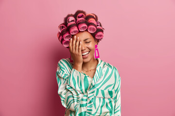 Funny joyful woman makes face palm, laughs at something, wears hair curlers, makes perfect...