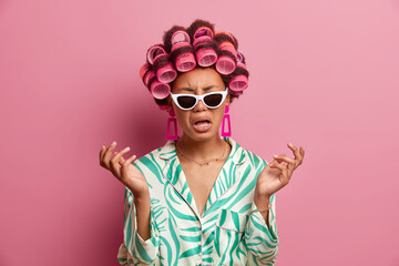 Frustrated dark skinned young woman being in bad mood, expresses negative emotions, raises hands with disappointment, feels bored and lonely, wears trendy sunglasses and hair curlers on head