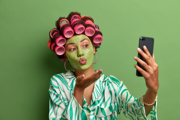 Portrait of attractive woman takes selfie, sends air kiss at camera of smart phone, has romantic mood, makes photo for husband, applies green nourishing mask on face, wears hair rolles, poses indoor