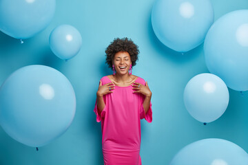 Fototapeta premium Indoor shot of cheerful curly woman in pink fashionable dress, closes eyes, prepares for speical occasion, glad to recieve congratulations with birthday, isolated on blue backgroud, inflated balloons