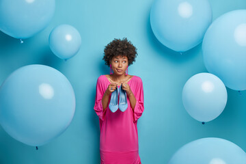 Fototapeta premium Pensive indecisive woman thinks what wear on foot to fit dress, holds high heeled blue shoes, dresses for birthday party, wears fancy long pink dress, isolated over studio wall, air balloons