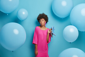 Fototapeta premium Lovely dark skinned curly African American woman keeps lips rounded, dressed stylish dress, holds shoes, chooses what to wear or dress, isolated on blue background with inflated balloons around.
