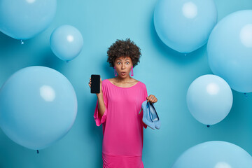 Photo of astonished curly young woman shows mobile phone display and high heeled shoes, makes shopping online, buys clothes in internet store, stands against blue background with balloons around