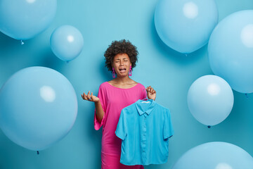 Fototapeta premium Womens fashion collection concept. Depressed crying dark skinned woman organizes clothes in wardrobe, holds blue shirt on hangers, has bad mood, thinks what to wear for party, blue background