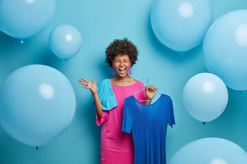 Fototapeta premium Emotional overjoyed dark skinned lady happy to have occasion for wearing favorite outfit, poses with long blue dress on hanger, has excellent mood, enjoys celebration and holiday preparation. Fashion
