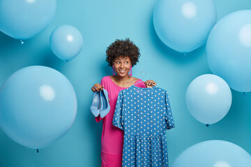 Fototapeta premium Style, clothes concept. Fashionable woman buys outfit for special occasion, waits party, holds fancy polka dot dress on hangers and blue high heeled shoes, surrounded by big inflated balloons