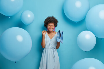 Fototapeta premium Women, fashion, clothes concept. Glad young African American woman clenches fist with joy, rejoices new purchase, buys fashionable outfit and shoes to dress for special occasion, blue color prevails