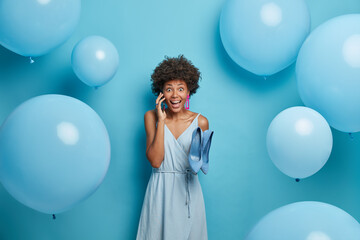 Fototapeta premium Beautiful winsome joyous woman organizes and prepares party event, invites friends via smart phone, chooses outfit to look brilliant, dressed in long dress and holds blue shoes, celebrates birthday