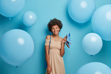 Happy elegant woman in stylish dress, carries blue bag on shoulder and heeled shoes in hand, poses against festive balloons, ready to celebrate something, prepares for party. Women and fashion concept