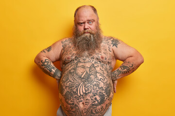 Serious bearded fat man has thick beard, tattooed body and big belly, looks from under eyebrows, keeps hands on waist, isolated on yellow background. Obesity, liposuction, weigh loss concept