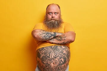 Serious surprised blue eyed man with thick beard, keeps arms folded, listens wife explanation,...