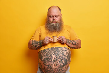 Serious bearded man with big tummy, tattooed arms and belly, holds very small carton cup of coffee...