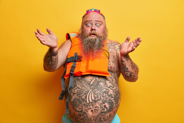 Indignant puzzled overweight man spreads hands sideways, has big tummy, cannot put on safety vest, wears swimming goggles, has summer recreation, isolated over yellow background. Water activities