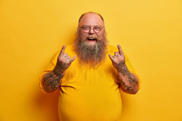 Obese funny man in yellow t shirt, shows heavy metal sign, attends concert of favorite music band,...