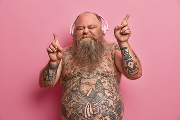Relaxed funny thick man with naked body, tattooed arms and stomach, dances while listens music, moves arms and closes eyes in enjoyment, wears headphones on ears, has fun and feels aspiration