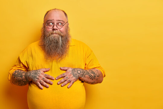 Pensive bearded bald man keeps hands on big tummy, stands in thoughtful pose, has tattooed arms, thick beard, wears round glasses, isolated over yellow background, blank space aside, thinks or doubts