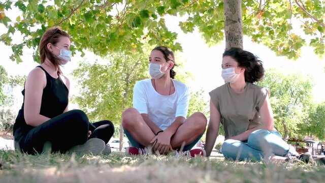 Young friends talking and laughing while sitting on grass. Meeting with friends in the park during the coronavirus epidemic. Following the rules of social distancing and wearing face masks.