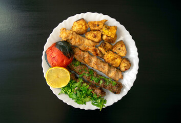 Famous traditional Arabic, Turkish, Israel food. Grilled chicken shashlik, lamb, beef kofta kebab, tomato, parsley, lemon on white plate on dark wooden background, top view. Kafta kebab