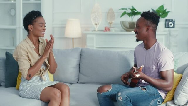 Young talented afro-american man singing song with ukulele guitar for his woman. Excited happy girlfriend hugging her man. Romantic date. Entertainment.