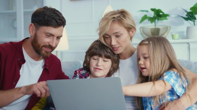 Affectionate cute caucasian family with young parents and happy two children watching funny internet videos on laptop enyoing leisure time together.