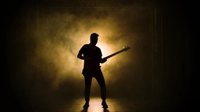 Silhouette of a young guy playing on the electric guitar on stage in a dark studio with smoke and neon lighting. Slow motion.