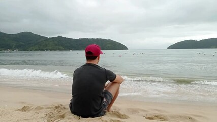 44 year old Brazilian man sitting with his back on the beach sand, watching the sea.