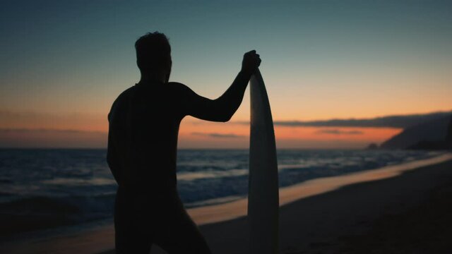 Silhouette of surfer standing with board and watching sunset, adjusting hair, sunset over the ocean, coast of Pacific ocean