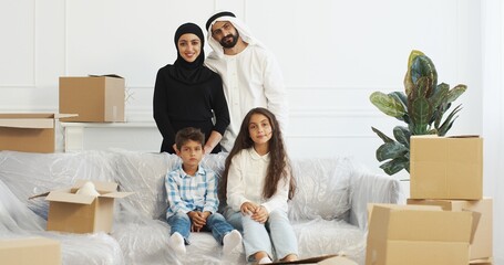 Portrait of happy muslim family smiling to camera and posing in living room. Moving in new accommodation. Arabian mother, father, daughter and son among carton boxes at home.