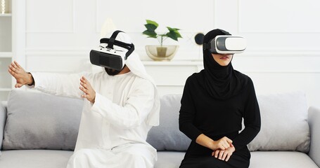 Arabic man and woman in traditional emirates clothes in VR glasses sitting on couch at home and...