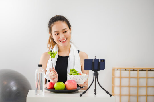Beautiful asian woman healthy blogger is showing apple fruit and clean diet food. In front of smartphone to recording vlog video live streaming at home.Fitness influencer on social media online.