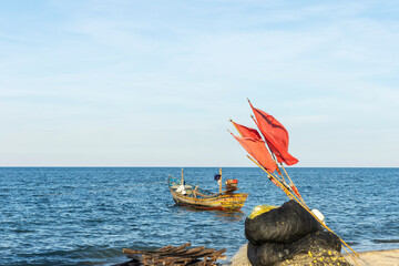 Small local fishing boat parks next to a clean beach against light waves under bright blue cloudy sky. Foreground of red flags for fishing