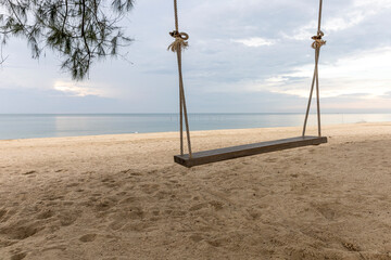 Fototapeta premium Little wooden swing under shade of big trees at peaceful beach on late afternoon. Blue cloudy sky and calm sea