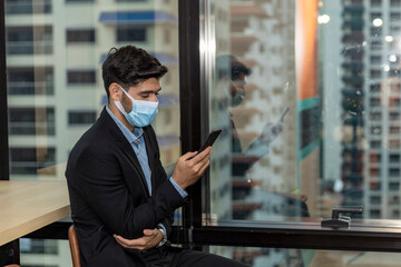 Young CEO or businessman with surgical face mask using mobile phone during office hours as part of Business New Normal and lifestyle during COVID-19 or Coronavirus outbreak. Man wearing face mask