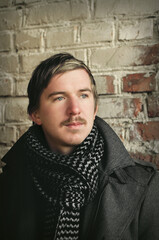 portrait of a young mustachioed guy in a warm coat and scarf against an old brick wall