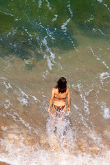 Aerial image of a beautiful long haired young caucasian girl wearing bikini is walking into the sea from the beach. It is a hot summer day. Water splashes around her as she walks into the water.