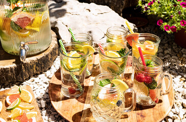 Refreshments with lemon, lime, orange, grapefruit and mint in glasses and a large jug with a crane stand on a wooden tray in the open air in the garden. Beautifully designed catering