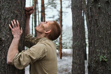 young soldier with mud and bruises on his face leaned against the trunk of a pine tree in the winter forest