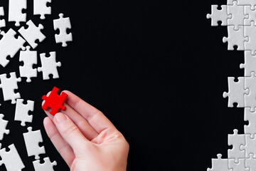 Female hand is holding red piece. White puzzles are scattered on left side and stacked on right. Process of structuring. Becoming like everyone else in global state system. Fight against individuality