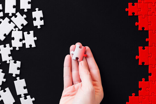 Female hand is holding piece colored in gradient. White puzzles are scattered on left side and red stacked on right. Process of transformation. Becoming like everyone else in global state system.