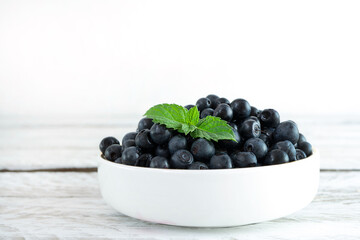 A white bowl full of juicy blueberries stands on a light wooden background. The bowl is decorated with fresh mint leaves. Delicious seasonal berries, a healthy dessert. Close-up
