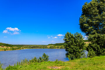 Obraz premium Summer landscape with beautiful lake, green meadows, hills, trees and blue sky