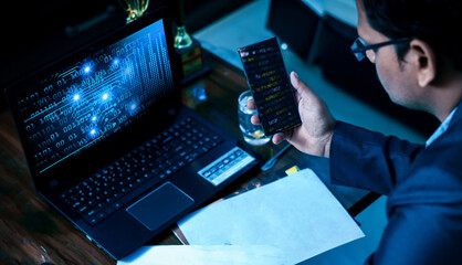 Anonymous hacker programmer uses a laptop and smartphone to hack the system in the dark. Concept of...
