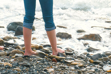 Woman stands on a stone beach in the waves. Feet close-up