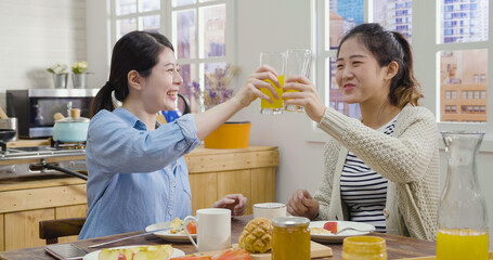 relax asian japanese female friends clink glasses in home kitchen during breakfast. two beautiful women drink fresh orange juice in morning house. wooden table with delicious healthy dishes.