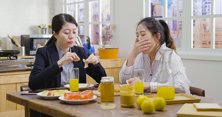 stylish tired woman in sleepwear yawning while business woman roommate have breakfast sitting beside. exhausted female in pajamas wake up in early morning and cover mouth with hand. lady eat bread